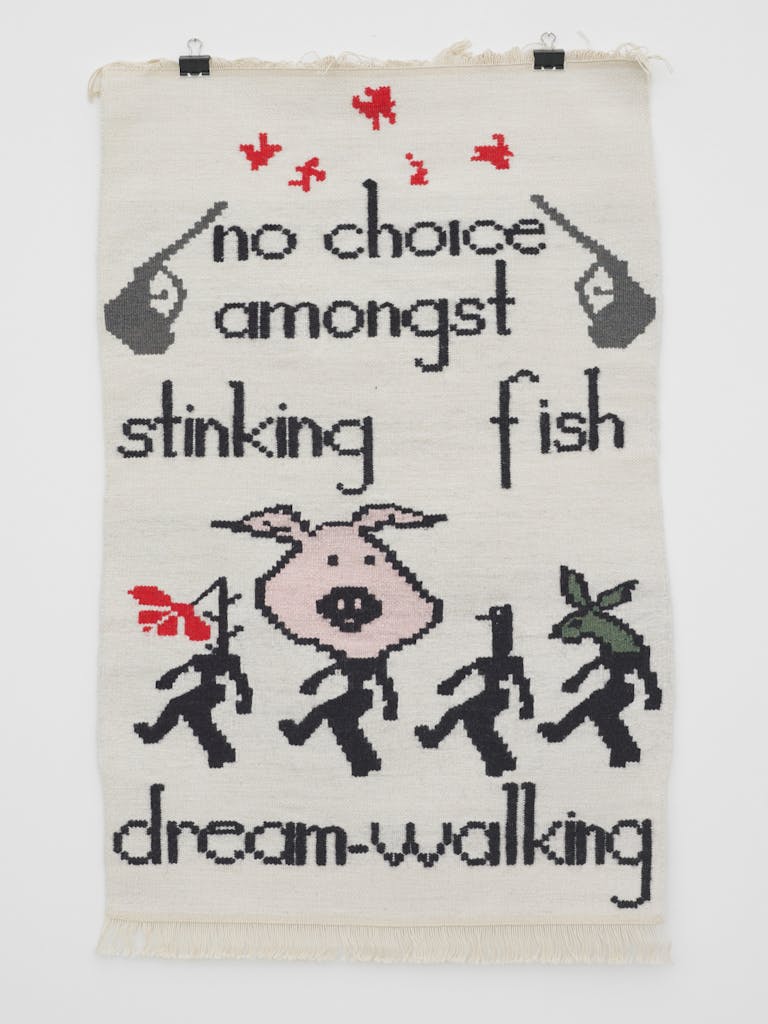 A photo of a cream tapestry in which the title of the work is woven alongside blocky images of guns and marching figures, one of which has a pig's head