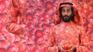 A man with a surprised expression holds a pomegranate in two ands; his clothing, as well as the background, is printed with more pomegranates