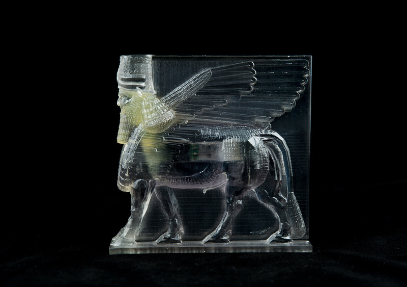 A transparent 3D printed ancient artifact, depicting a winged lion with the head of a bearded man, set against a black background