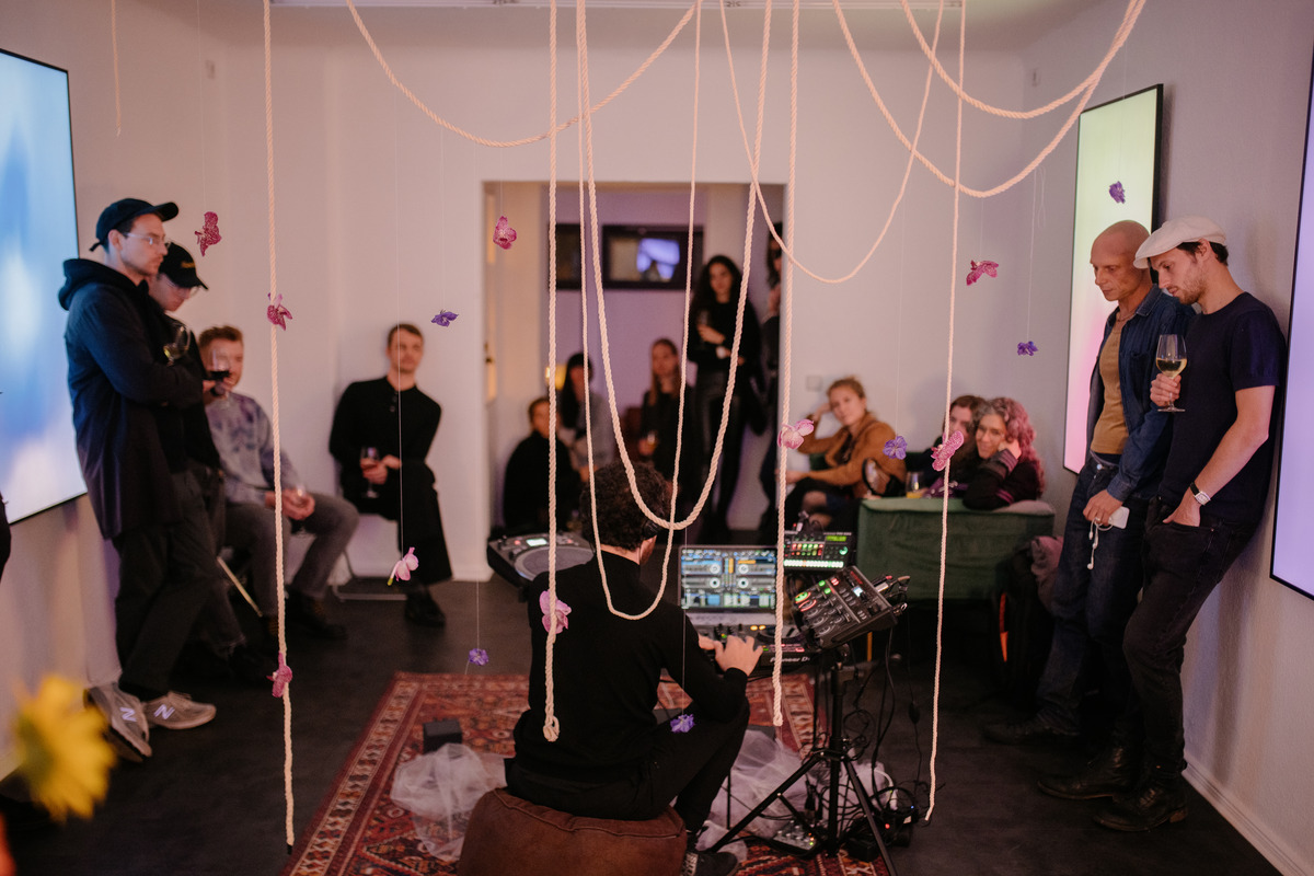 People stand in a room around someone making music with a computer and a mixer. Strands of pink thread with flowers attached dangle in the foreground