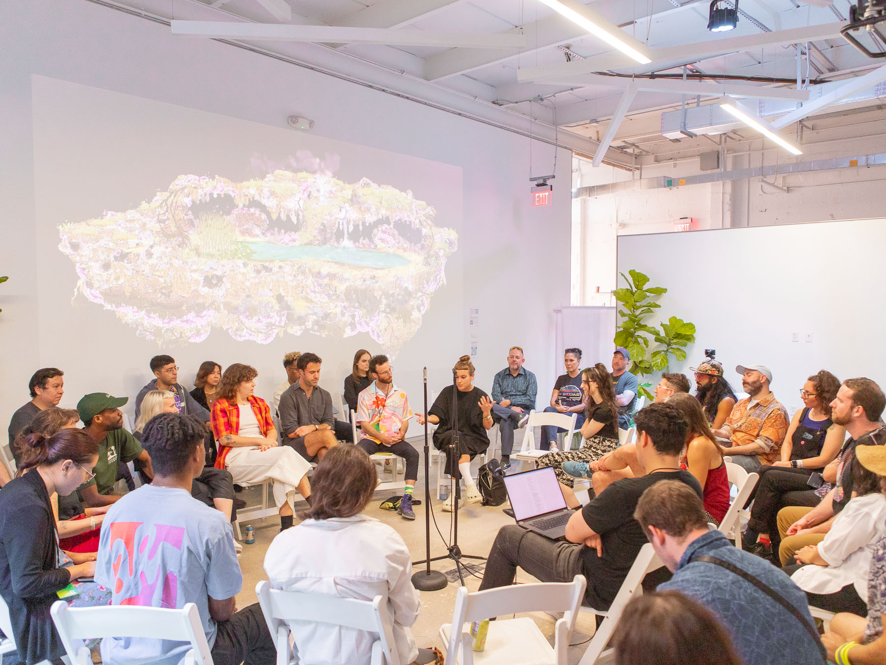 People sit in a circle of white folding chairs, listening to a person seated in the middle who is speaking and gesticulating. Digital art is projected on one of the walls