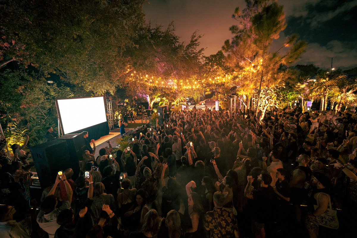 A crowd of people stand at an outdoor concert at night. THe venue is surrounded by green trees, with some yellow lights strung between them