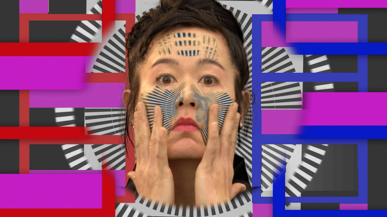 A close-up of a woman's face surrounded by boldly colored abstract shapes resembling a test pattern on a screen. Her eyes are opened wide and her fingers touch her cheeks, seeming to spread radial black and white lines across them