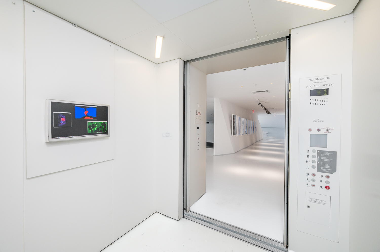 A photo of a white-painted elevator with a work on a monitor on view on one wall and a view into the corridor beyond