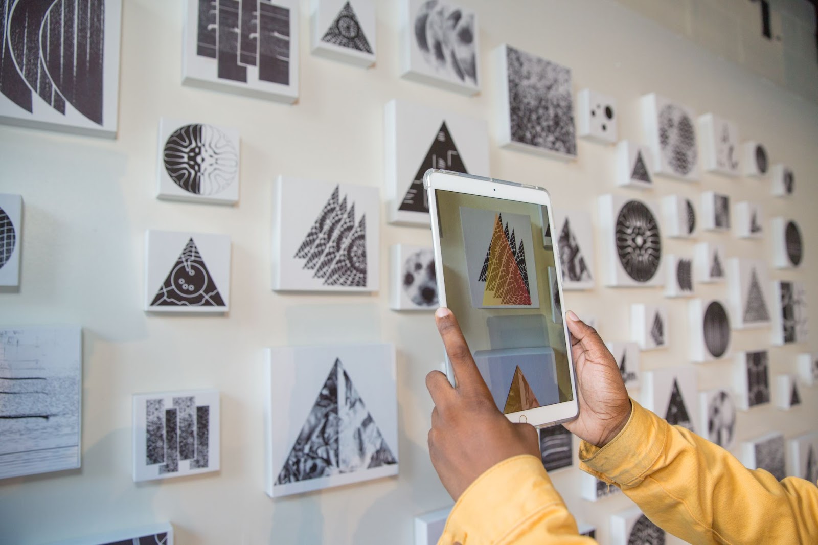 A photo of wall of small black and white geometric prints with the hands of a person holding an electronic tablet in front of the wall