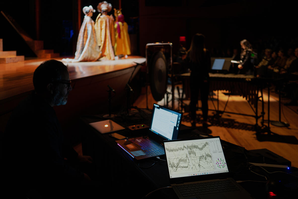 A photo of a darkened theater with people in fantastical costumes on a stage glimpsed in the background; a high-tech set-up with laptop and chart readings in the foreground