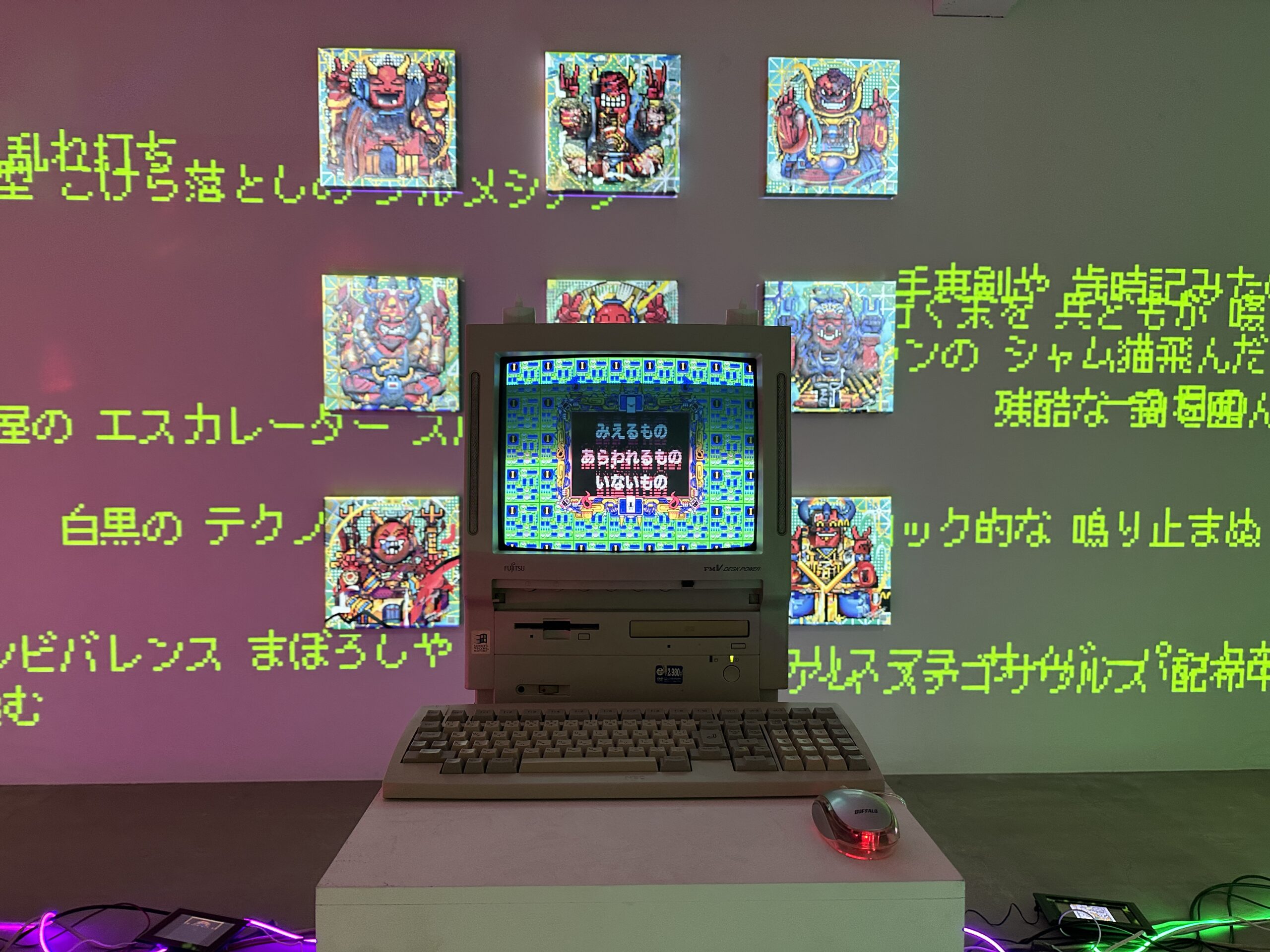 An old computer sits on a plinth in a gallery. Its screen displays 8-bit art with writing in Japanese; behind it, Japenese text is projected on the wall with green light, overlapping in places with prints of pixel art depicting folkloric creatures
