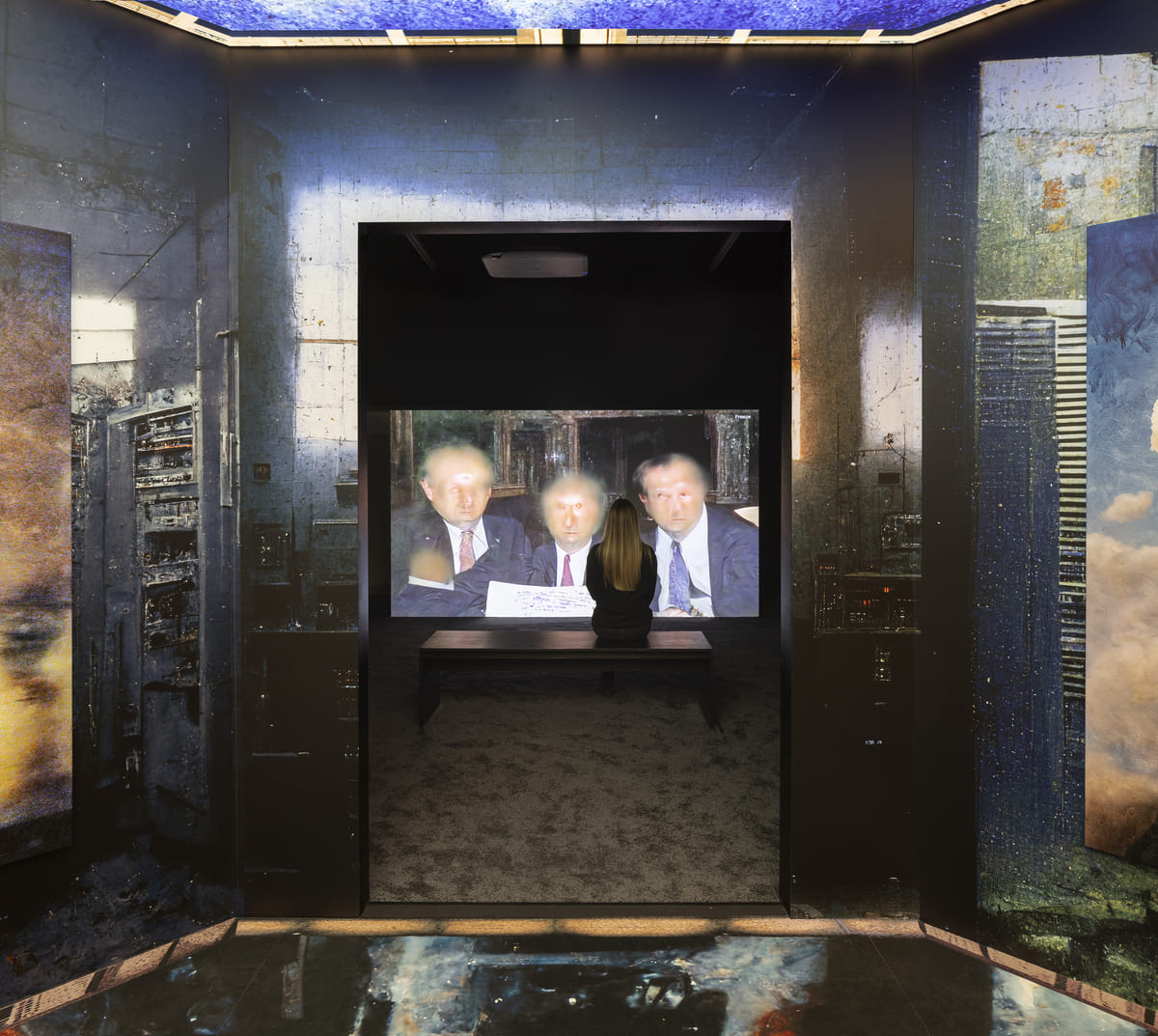 A photo of a gallery space in which a woman sits on a bench in front of a screen with a strange image of three blurry white men; she is viewed through a doorway wallpapered with an image of a dark cityscape