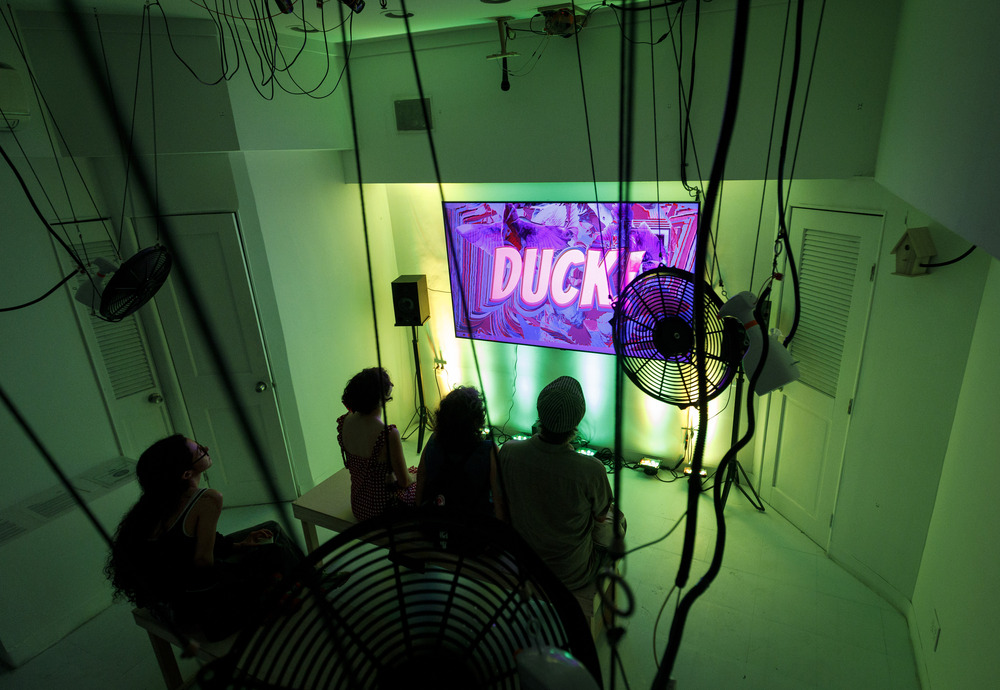Viewers sit on benches watching a video installation; the gallery is bathed in green light and fans blow on the room from above