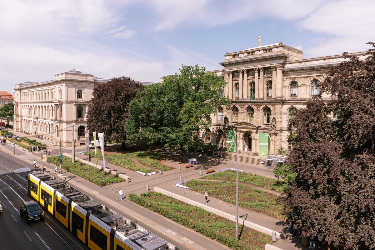 An aerial view of a grand stone building with a garden in the forecourt on a busy road, with a tram running across it