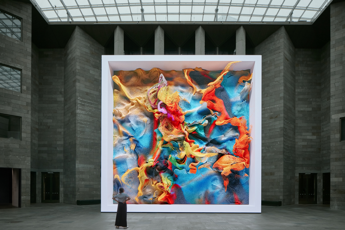 A large square screen with an abstract multicolored image seeming to pour out of it, with a woman in a blue dress standing in front of it