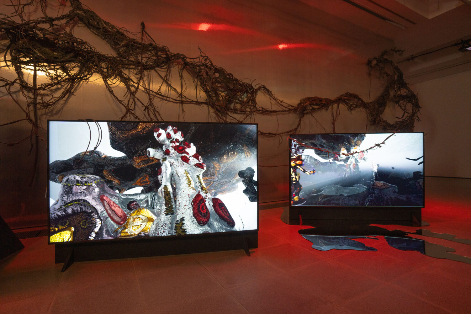 A pair of screens display fantastic landscapes. They are placed in a gallery where a vine-like sculpture hangs from the wall
