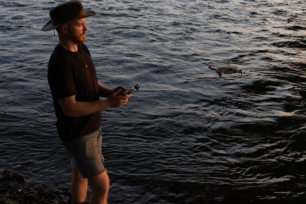 A man operates a drone beside a body of dark, swirling water