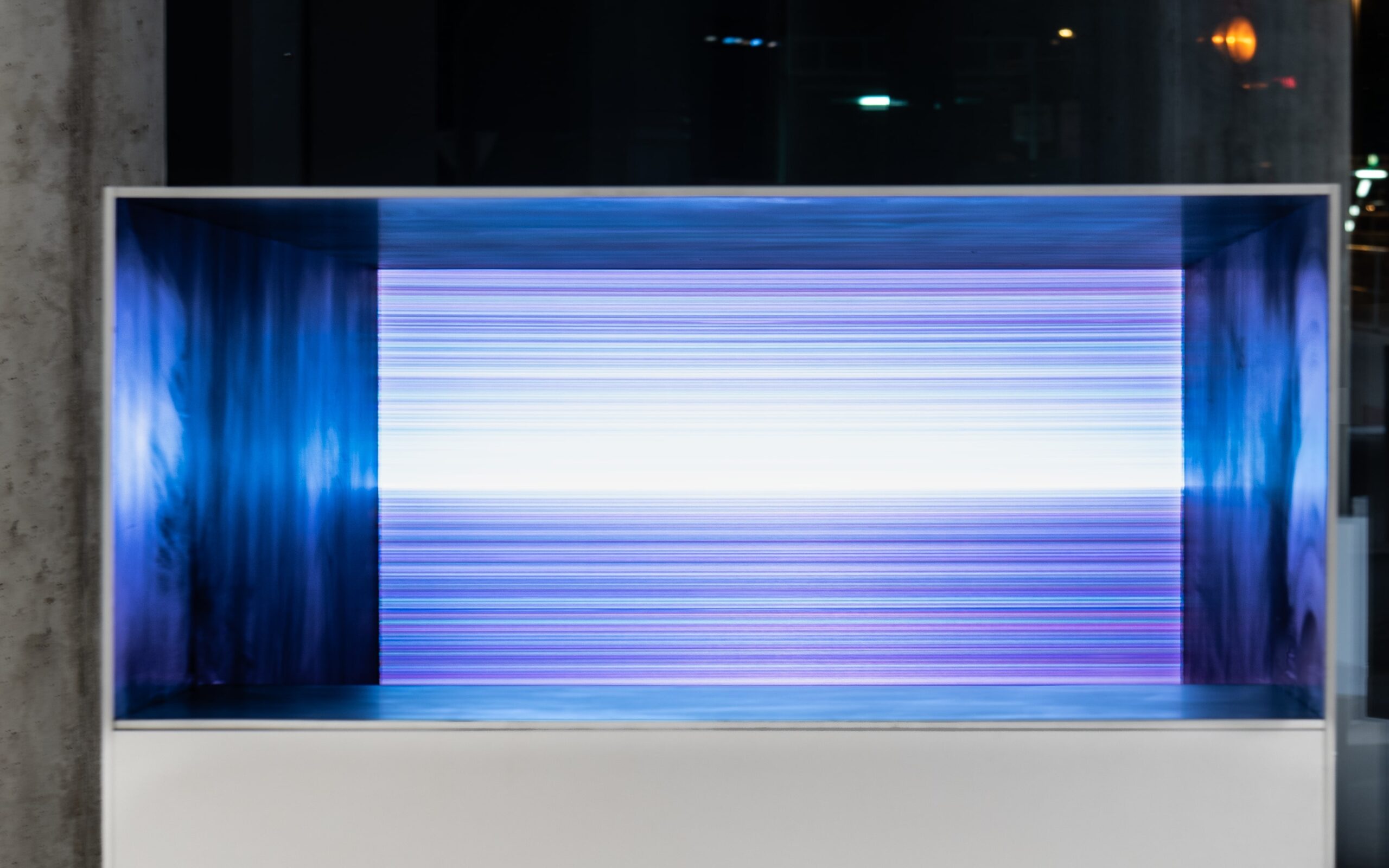 An installation view showing a futuristic box with a white base and filled with beams of blue and purple light