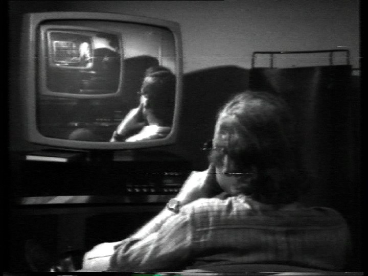 A black-and-white still of a man sitting in front of a TV