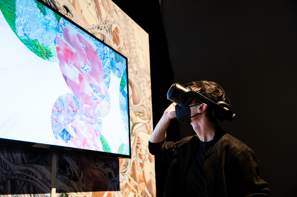A person in a VR headset stands in front of a screen that shows a view of the virtual art installation seen inside the headset; it feature pink, blue, and green spherical forms in a white void