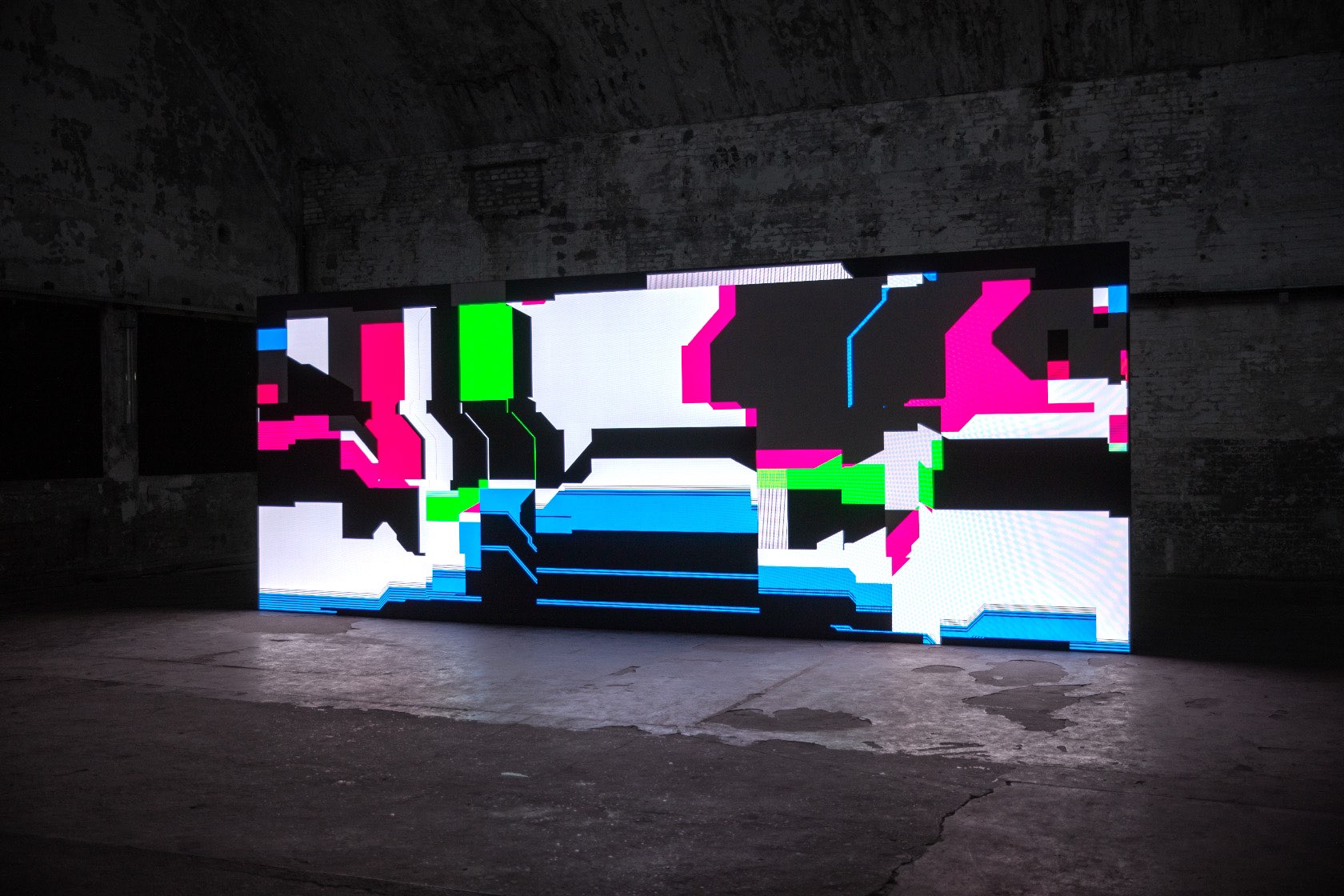 A digital work with shifting fields of white, black, pink, blue, and green is displayed on a large screen in a bare, concrete room