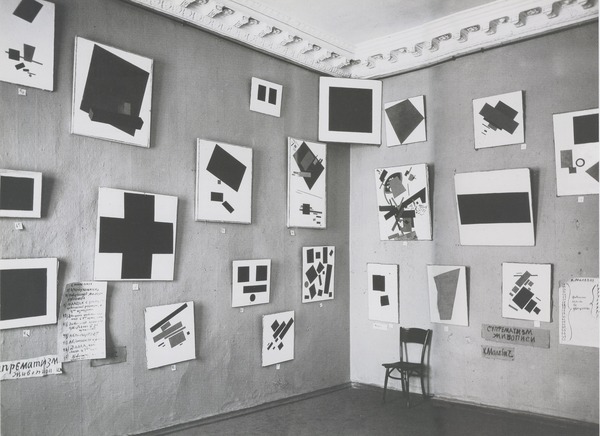 A black-and-white photograph of abstract paintings hung salon style in the corner of a room