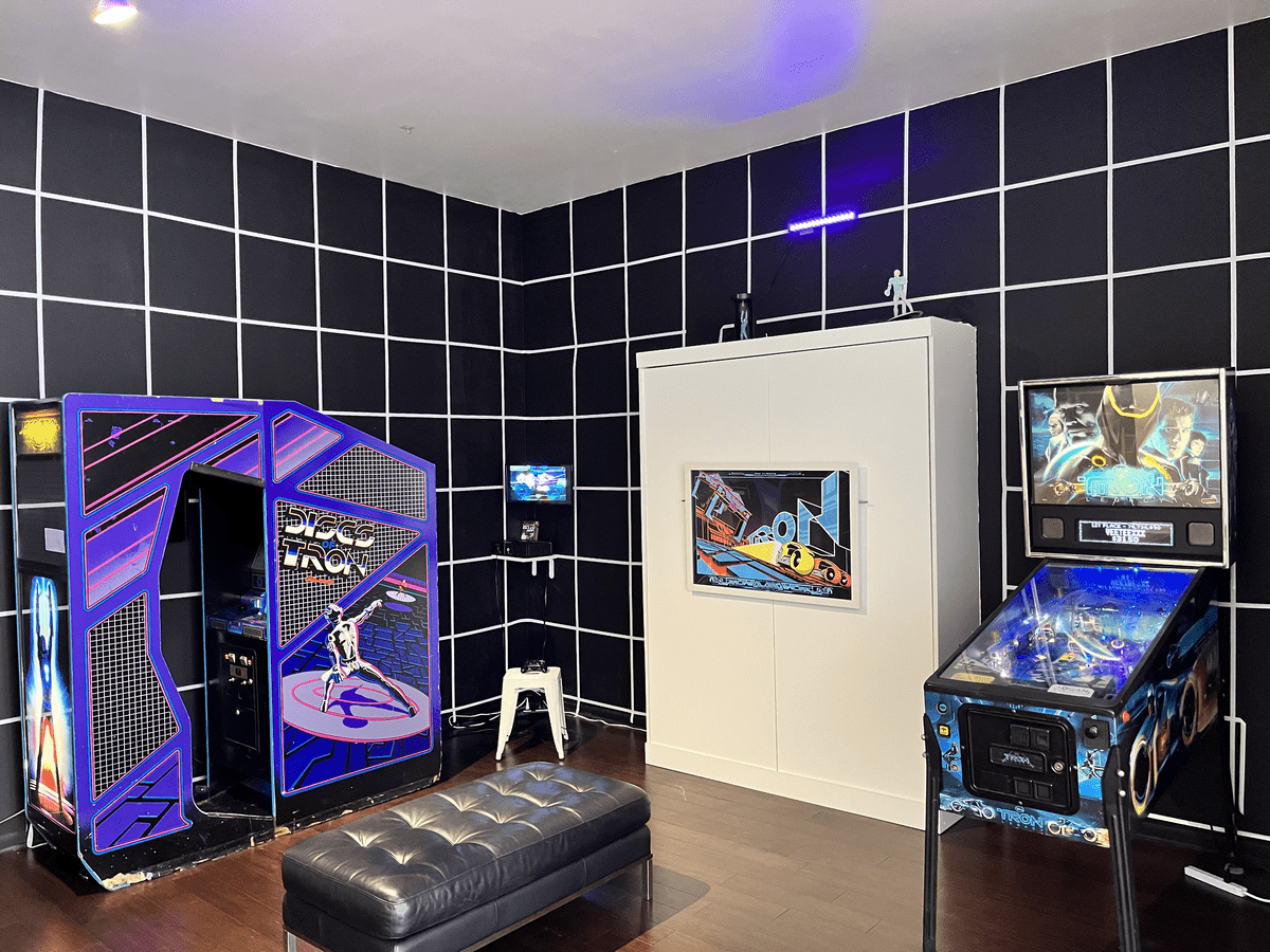 A photograph of an exhibition featuring an arcade game, a pinball game, and a console game on a small screen; the walls are painted black, with a white grid overlaid to enhance the designs of the game cabinets
