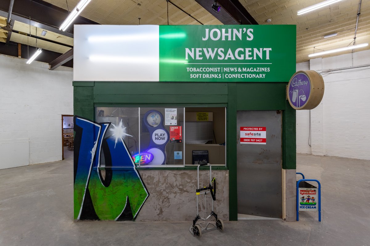 A photo of a conventional white cube art gallery containing a highly realistic replica of a shop called John's Newsagent