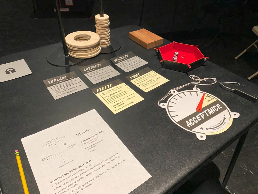 A photograph of a black table filled with game paraphernalia, including cards, a character sheet, dice, and wooden rings stacked on rods.