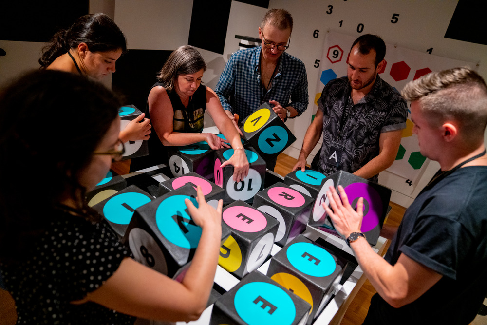 A photograph of people turning over cubes, the surfaces of which have black letters printed on pastel colored circles