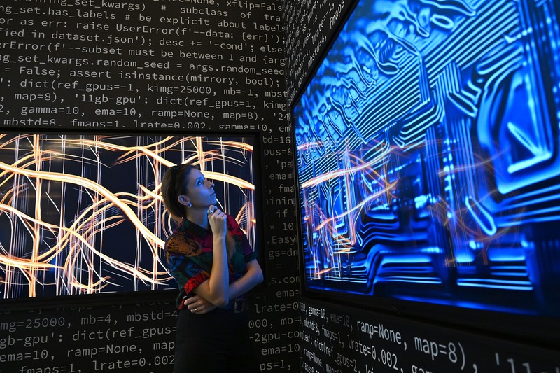 A woman contemplates abstract works recalling the contours of computer chips in a dark room where the walls are covered with lines of computer code