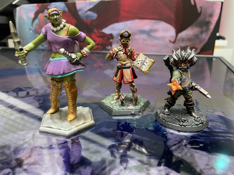 A photograph of three plastic statuettes representing fantasy characters: a tall female orc rogue, a magic user holding a spellbook, and an armored man with a thick blue moustache.