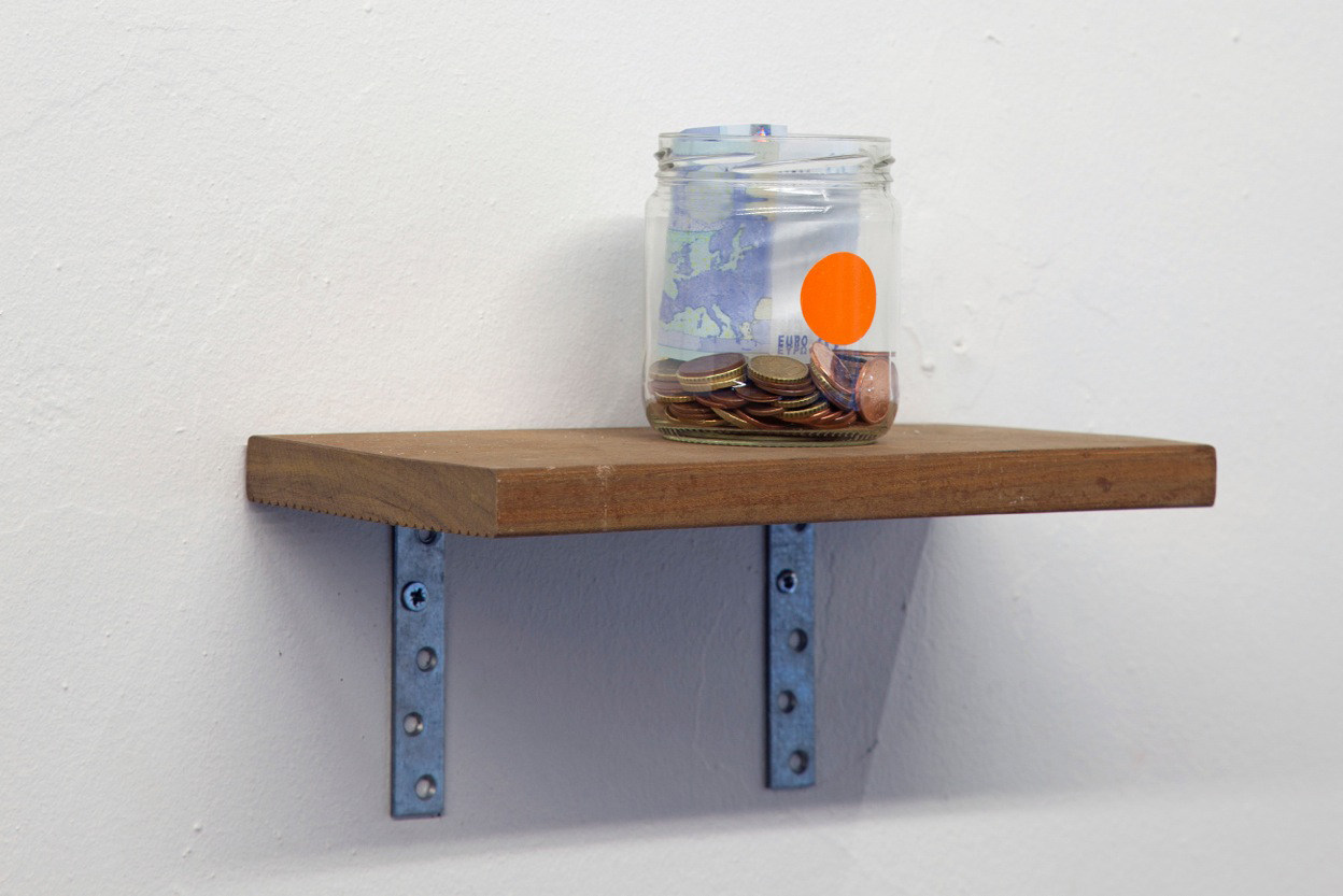 A jar filled less than a quarter to the top with coins sits on a modest wooden shelf