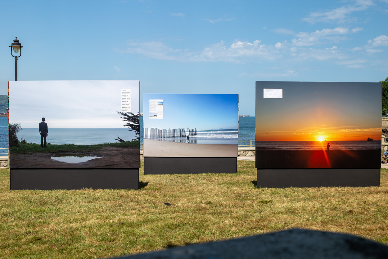 Large printouts of photographs of beach horizons, each with a dark silhouette of a person facing the ocean, stand on dark bases in a park at the edge of the water