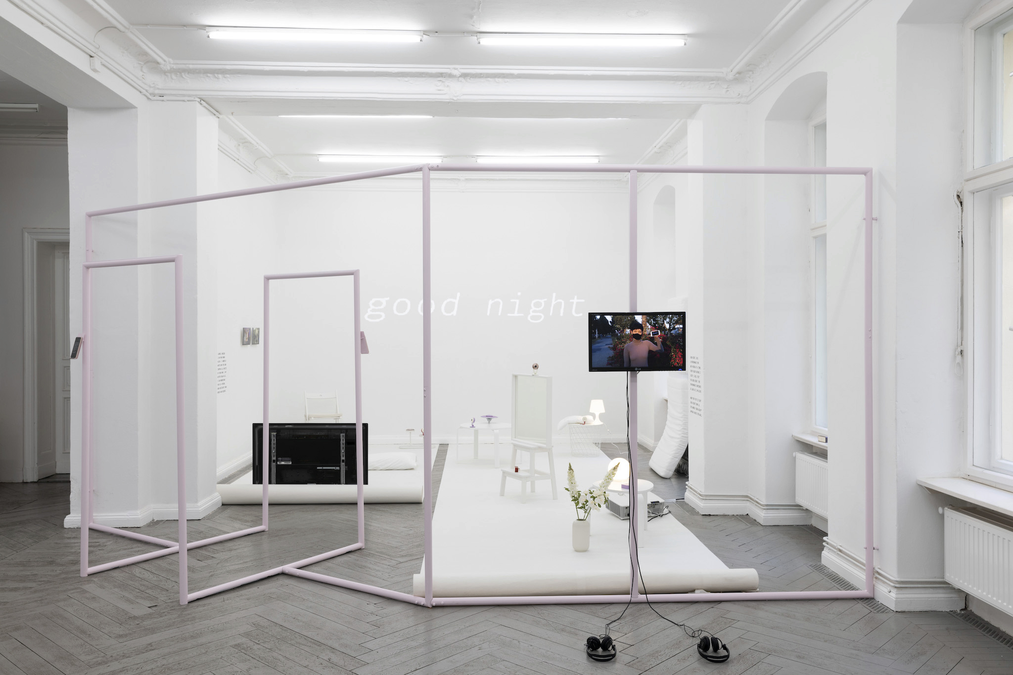 A photo of an installation of Lauren Lee McCarthy's work, involving a sculptural installation with monitors and a projection with the words "good night", in a white cube gallery