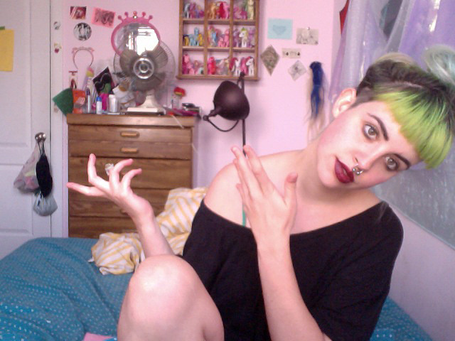A webcam image of a young woman with green hair sitting on her bed and gesturing to the pink wall behind her, which has a fan and cosmetics on a dresser, shelves crowded with figurines, and other domestic objects and mementos