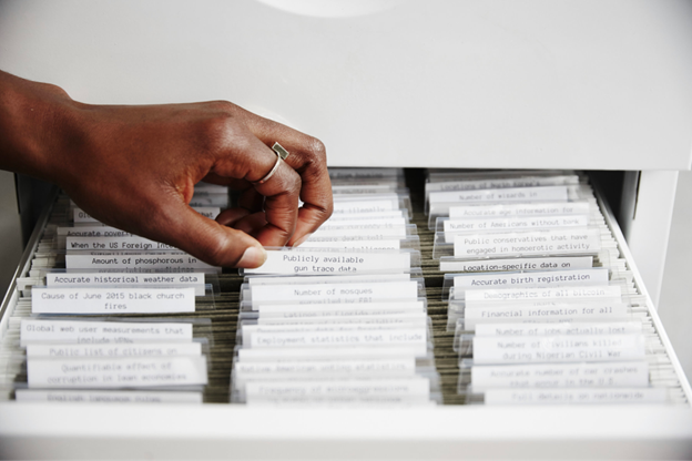 A photograph of a hand thumbing through file folders with labels like "Publicly available gun trace data" and "Cause of June 2015 black church fires"