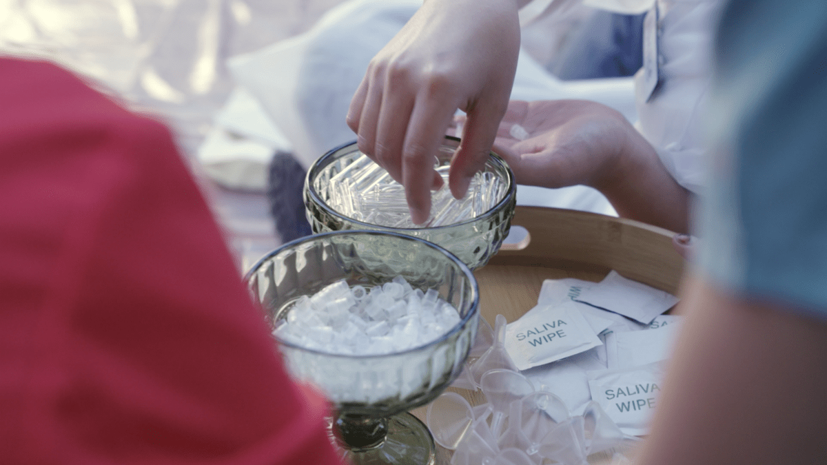 A photograph showing hands reaching into small bowls holding clear vials and stoppers. There is a pile of wet wipes labeled "SALIVA WIPE" on the table.
