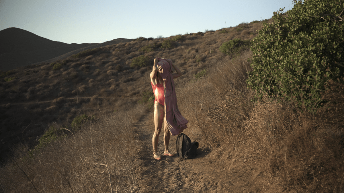 A photograph of a woman standing on a hillside, pulling a loose dress over herself while wearing a prosthetic pregnant belly