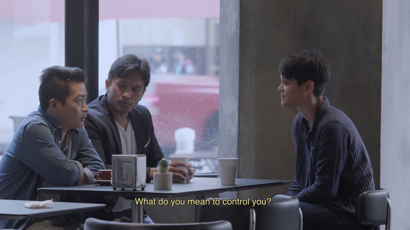 A video still of a gay couple speaking to a woman over a table at a coffeeshop. A subtitle reads: "What do you mean to control you?"