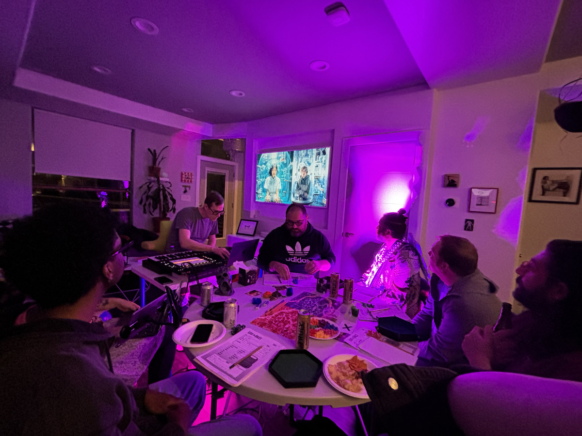A group of people is gathered around a table covered with snacks, drinks, gamepieces, and character sheets. The lighting is purple and atmospheric, and one attendee is operating a projected image via a laptop and sound with a mixing board