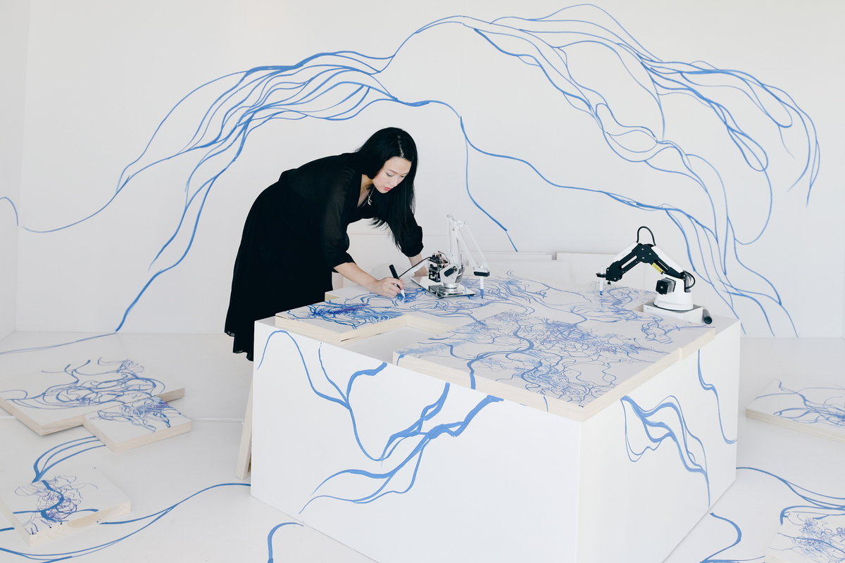 A photograph of a white room with a white canvas on a table on the center. A woman and a robotic arm are both drawing on the room and canvas with blue ink