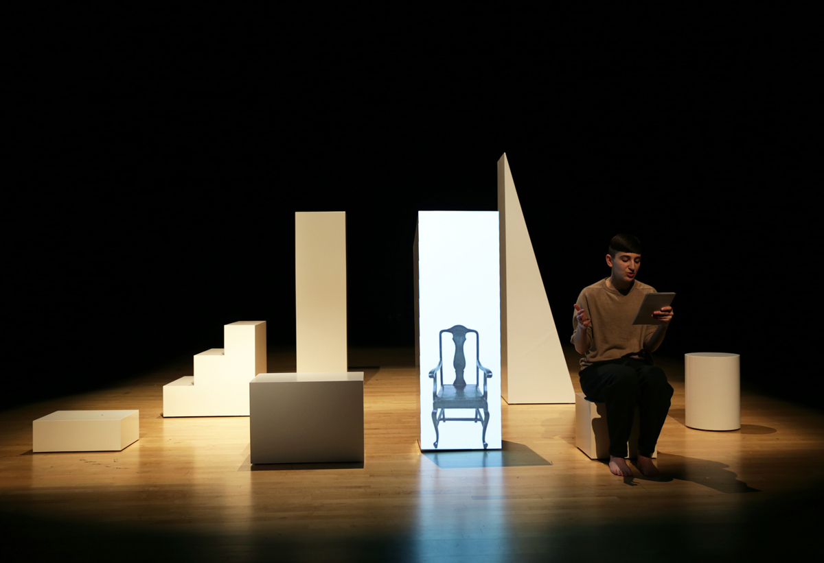 Photo of a spotlit stage in which a person sits on a white plinth. They are surrounded by other oddly shaped plinths, including one on which an image of an old-fashioned chair is projected