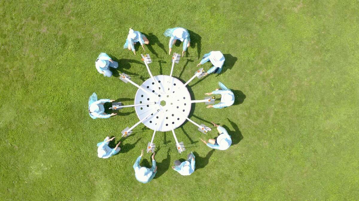 A birds-eye photograph showing ten people standing in a circle in a green field