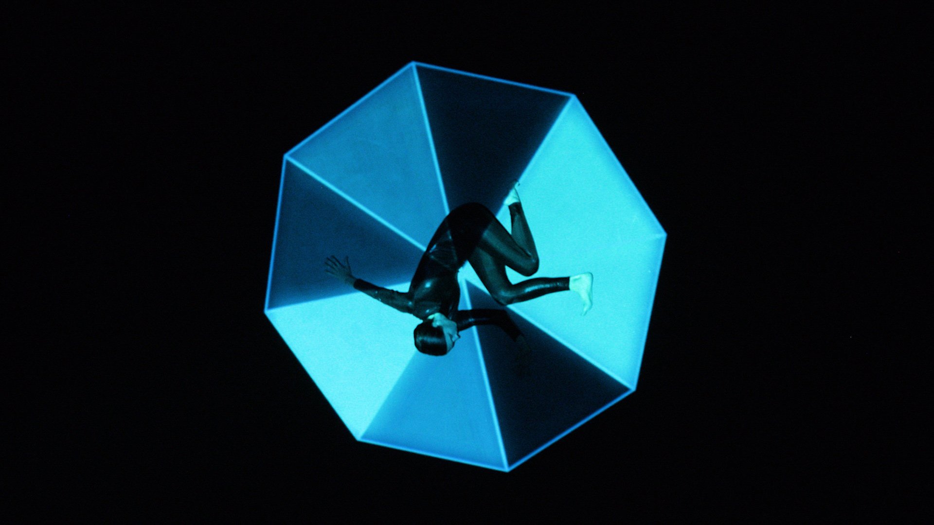 A projected image of a performer's prone body against an octagonal background of dark and pale blue shades that sides in a black void
