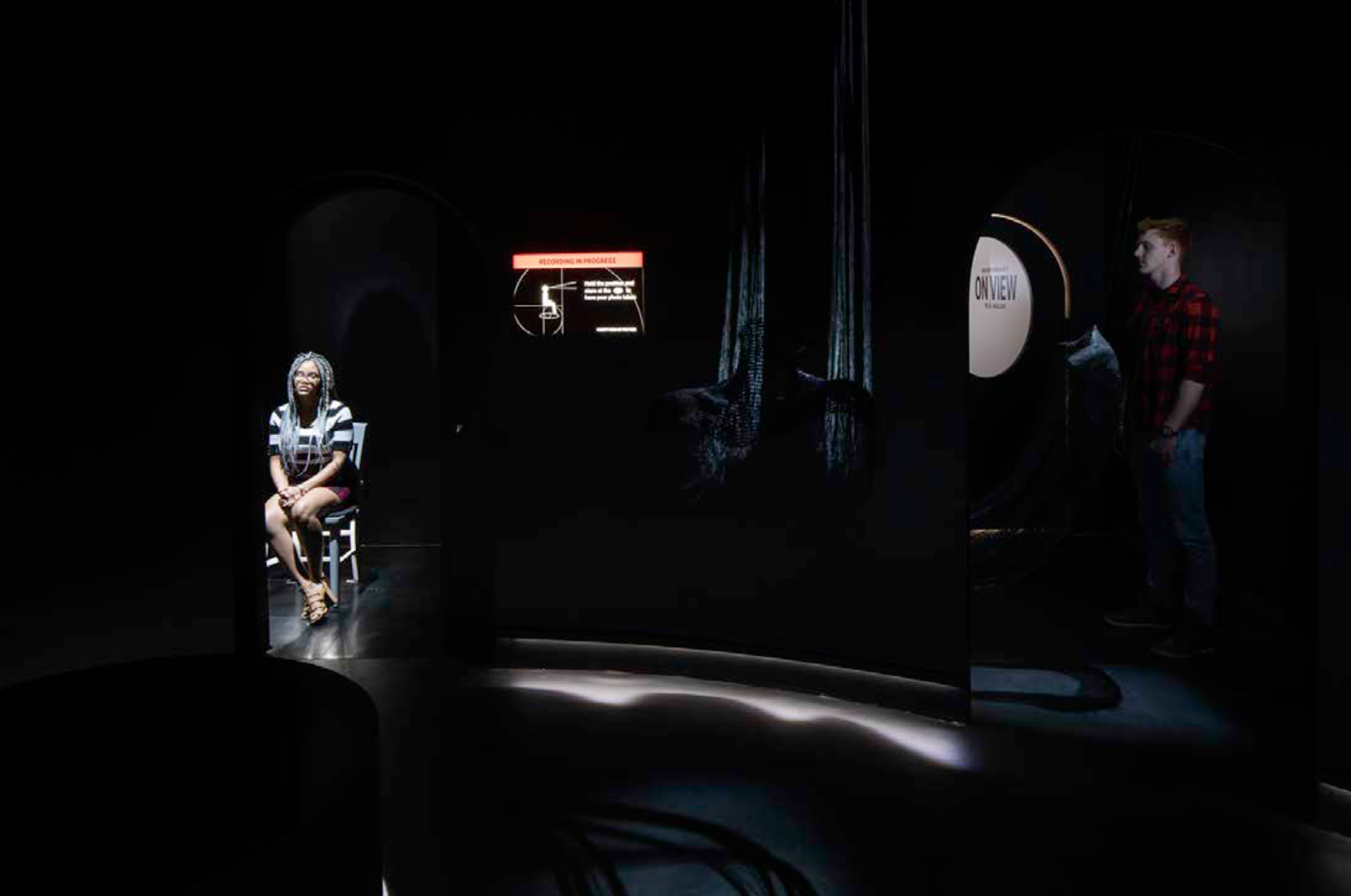 A photograph of a darkened space, on the left-hand side you can see a spotlit woman sitting in a chair, behind her there is a screen with text that reads "recording in progress". A man is visible on the far right of the image