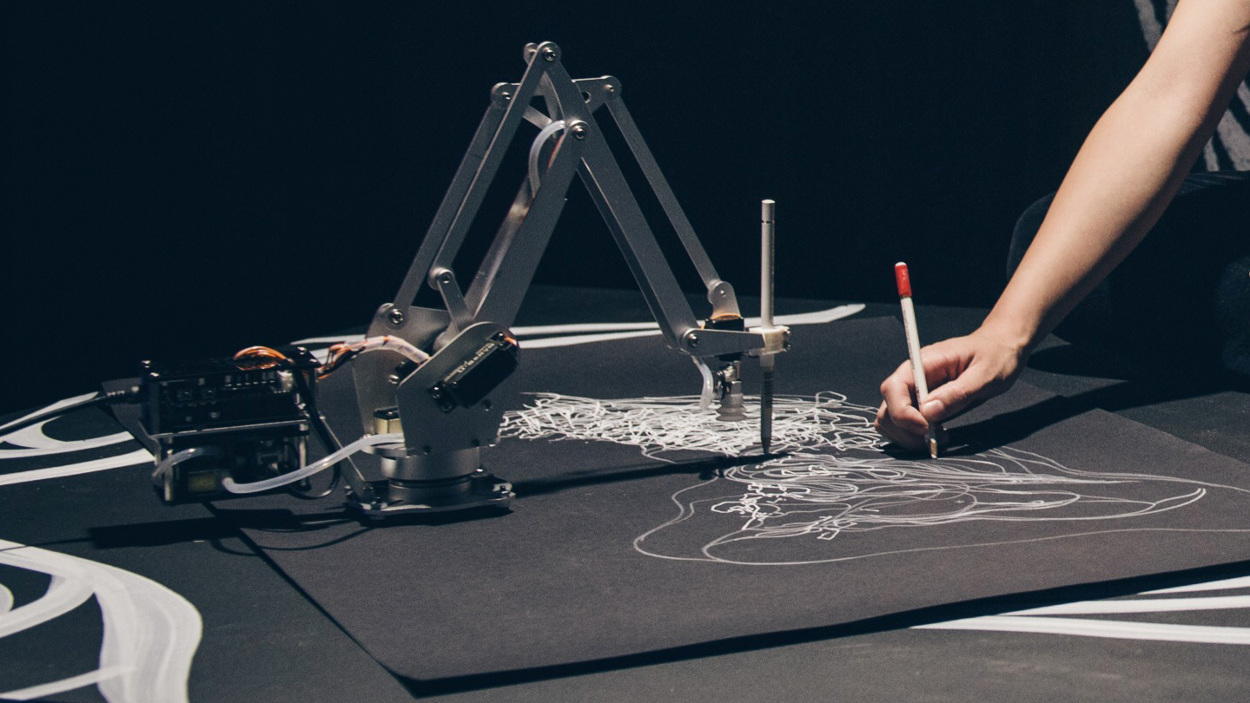 A photo showing the close-up of a woman's arm drawing on a black piece of paper alongside a robotic arm