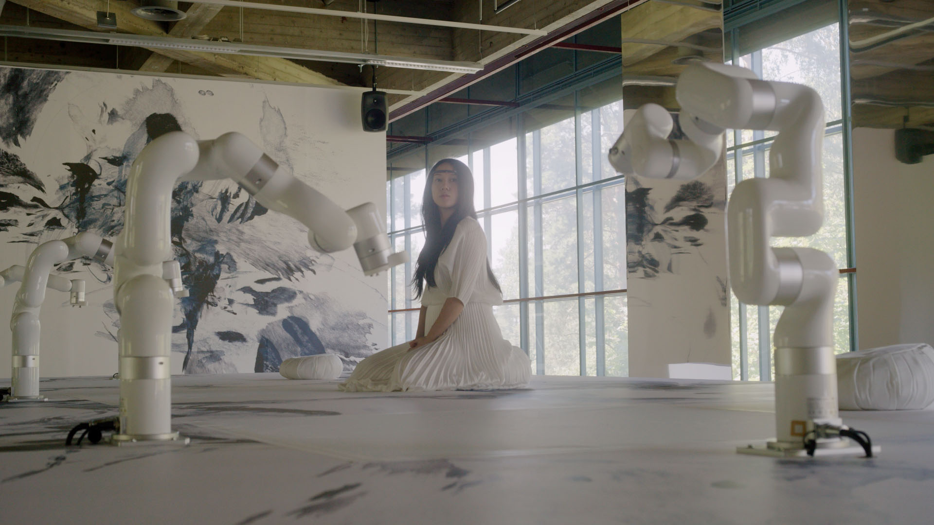 A photograph of a woman sitting on the ground, with some kind of electronic device around her head, surrounded by white robots and abstract drawings on the walls and floor
