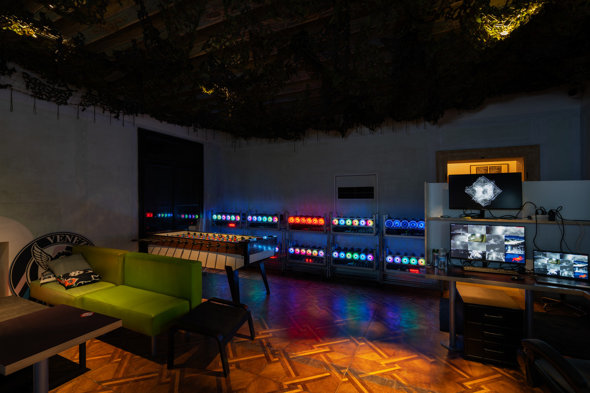 A photograph of a darkened room glowing with the rainbow light of many high-powered computers mining crypto currency, plus monitors displaying surveillance footage and action games