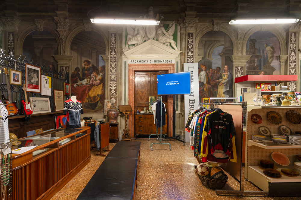 A photograph of a thrift store set up in an old church, with clothing and dishware on cheap shelves amid frescoes, under harsh fluorescent lighting