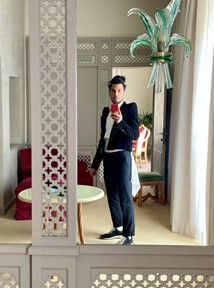 A photo taken with a phone by the subject in the mirror. He is standing in a large airy room with lots of natural light, wearing a stylish suit