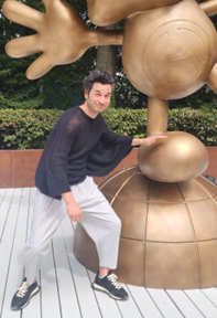 A photo of a man wearing a diaphonous black top with oversized sleeves. He is touching a bronze statue of a cartoon character and smirking
