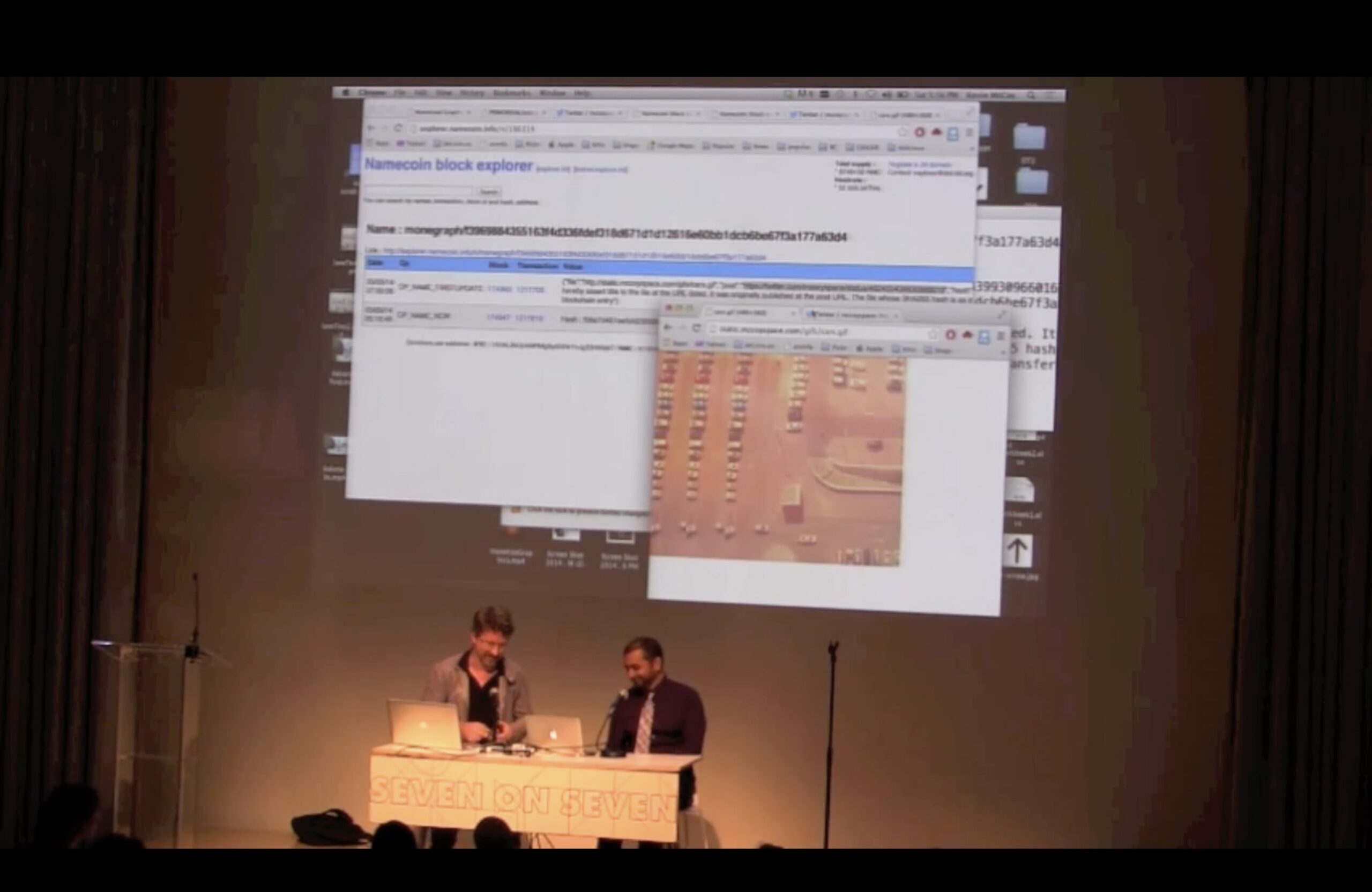 An image of two men sitting at a desk on stage with a presentation projected on the wall behind them