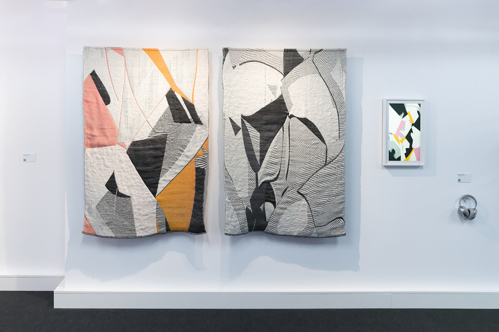 A photograph of two woven rugs hanging against a white wall. A small screen beside them shows an abstract image, similar to the design of the rugs